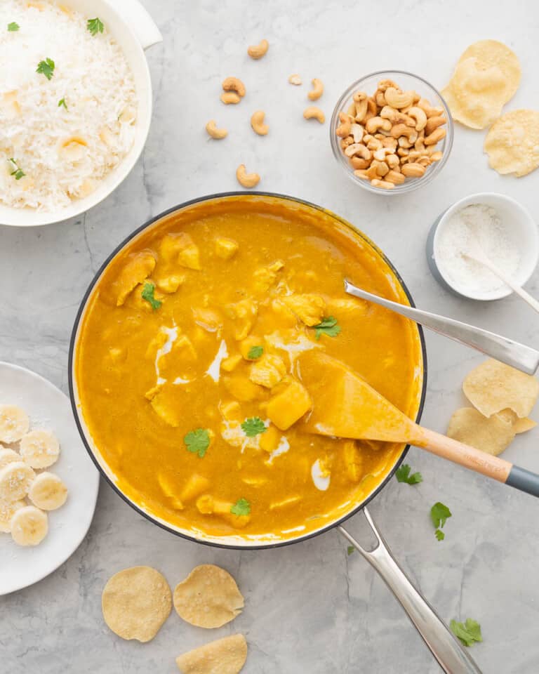 A large pan full of Mango Curry, drizzled with cream and sprinkled with parsley sitting on a bench next to a small bowl of cashew nuts, a ramekin of coconut, a bowl of rice and a plate of sliced bananas.