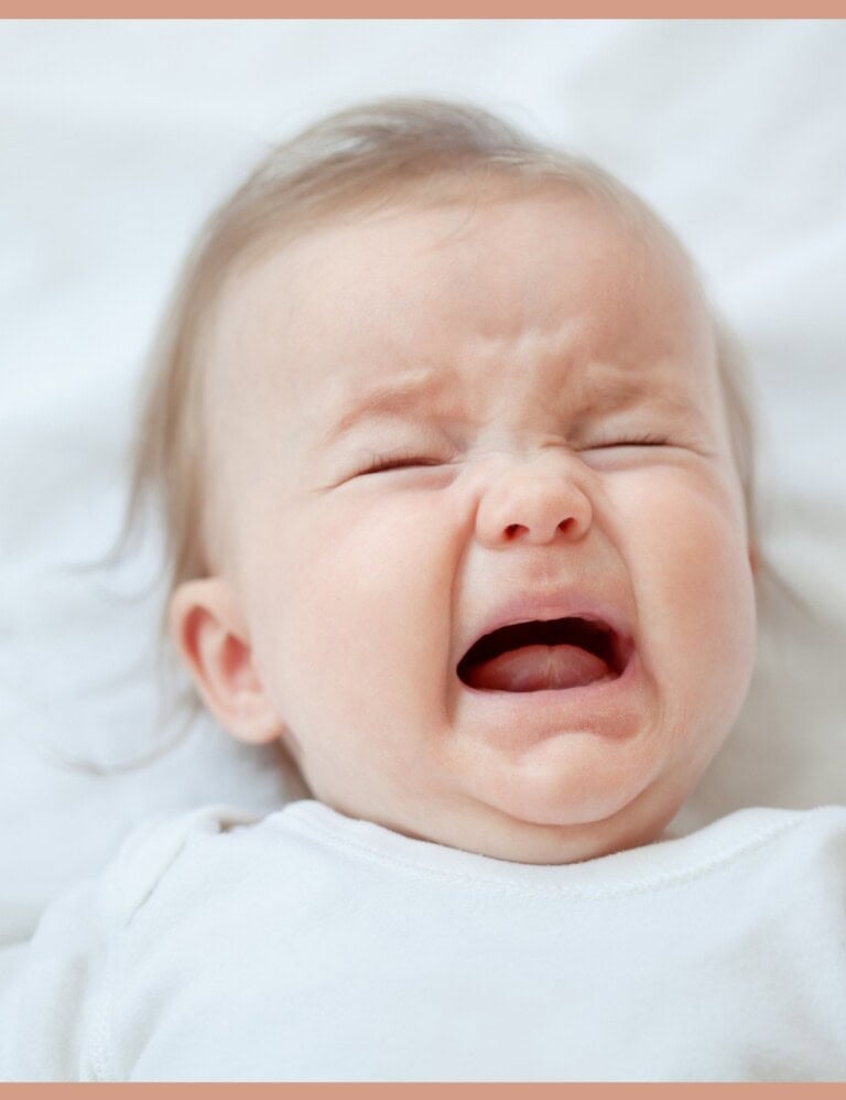 A crying baby in a white onesie on white crumpled sheets.