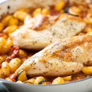 Two chicken berasts with crispy skin in a casserole dish with gnocchi and vegetables.