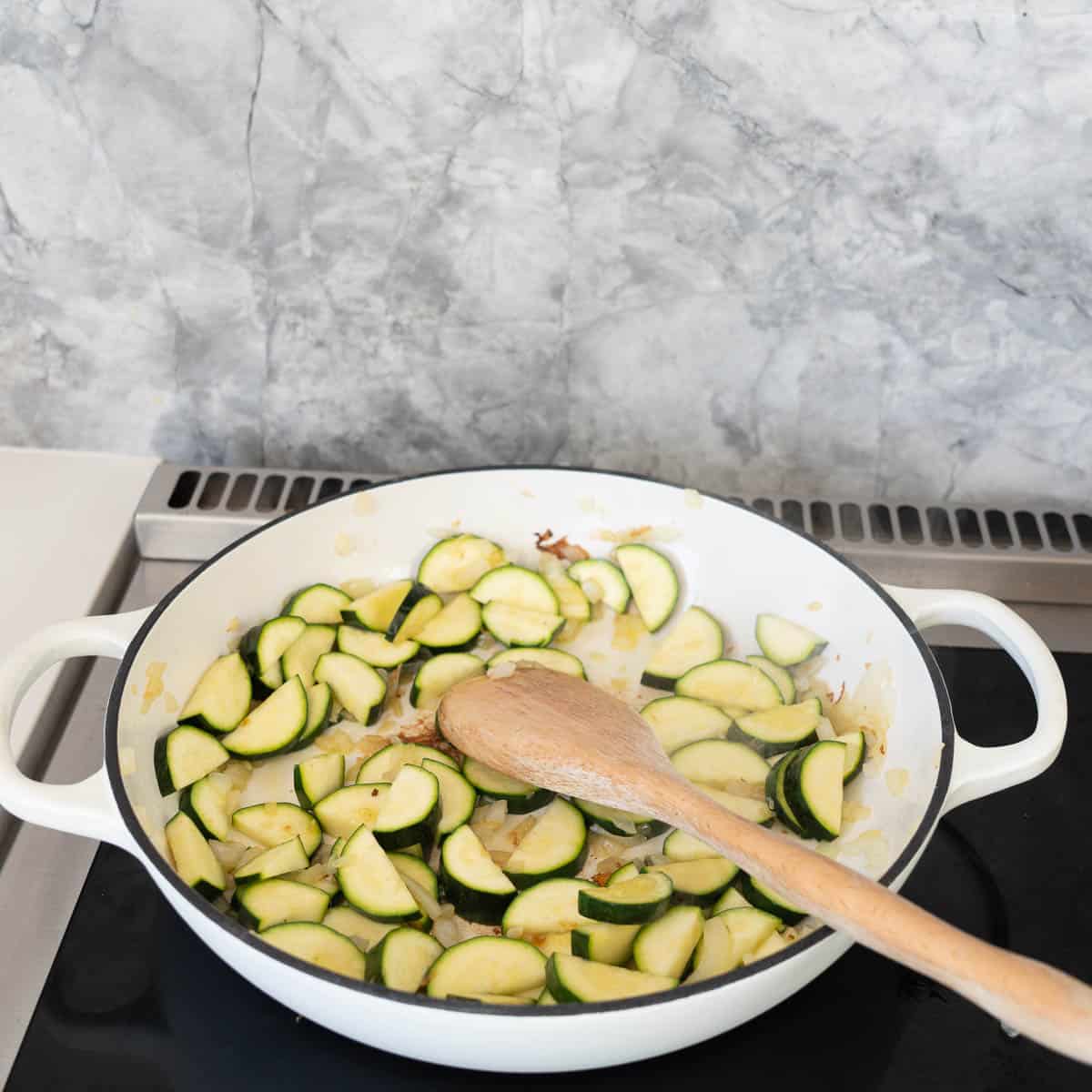 Slices of zucchini being pan fried in a large white casserole dish with a wooden spoon.
