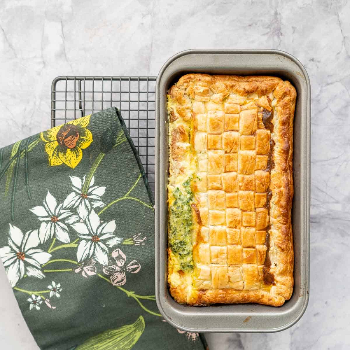 A pie with a golden brown lattice top in a loaf tin on a cooling rack.