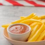 A bowl of fries with homemade ketchup in small ramekin inside bowl on benchtop with folded red and white stripped table cloth.
