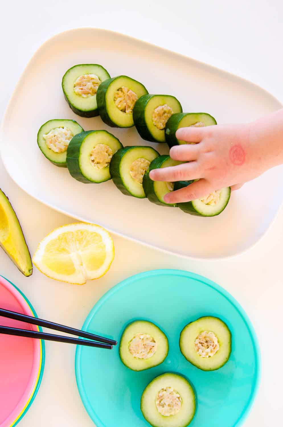 cucumber sushi on a white platter being reached for by a Childs hand, next to a stack of colourful kids plates.