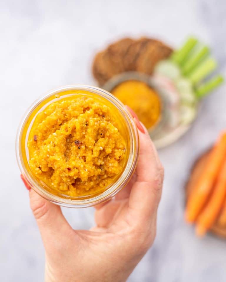 A jar of carrot pesto being held above a snack platter of vegetables and crackers.