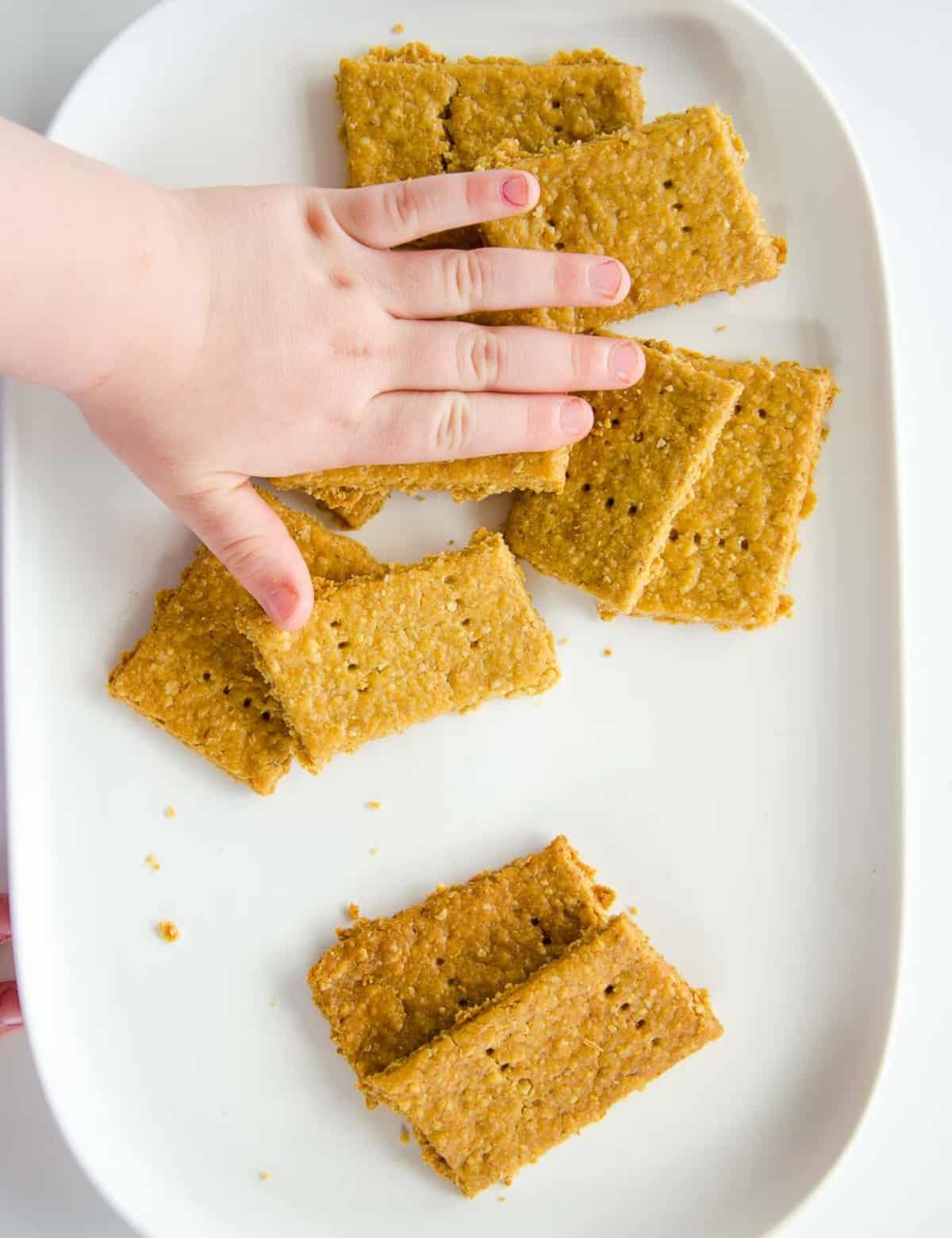 A toddler hand reaching for crackers on a white ceramic plate.