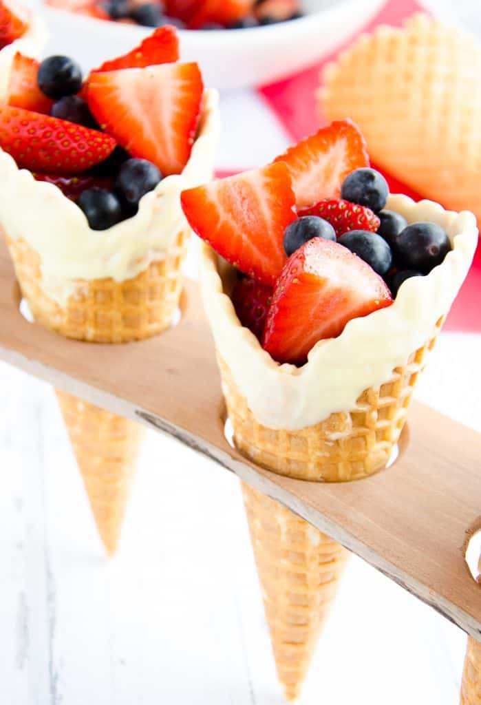 Waffle cone in holder, with white chocolate around the rim, with strawberries cut in half and blueberries inside.