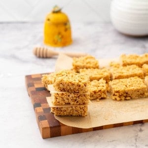 A tower of rice krispie treats on a wooden chopping board in front of a honey jar.