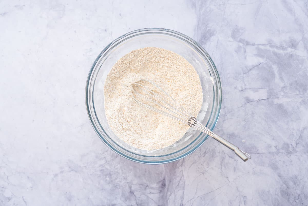 A glass mixing bowl filled with wholemeal flour with a whisk resting on the side.