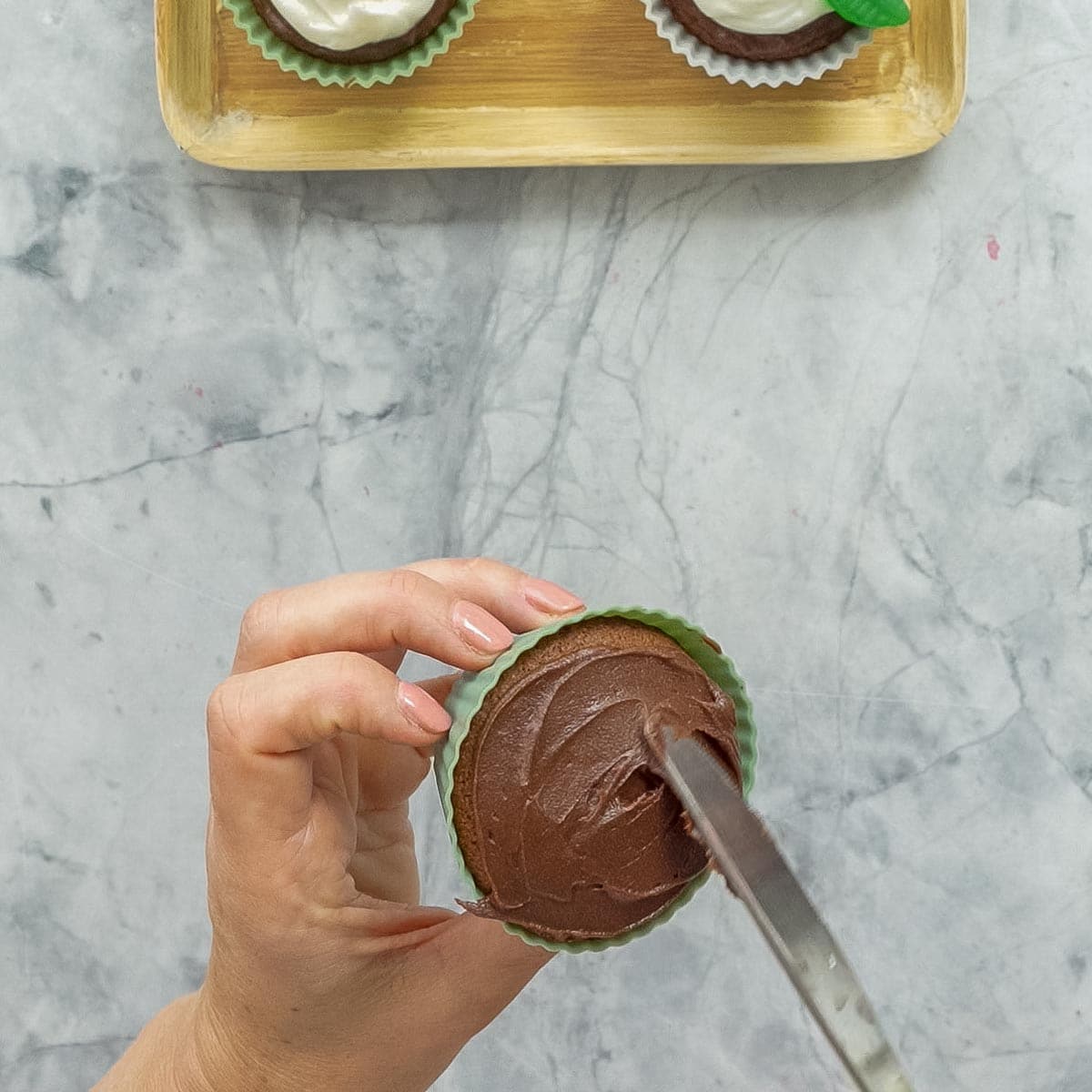 A chocolate cupcake being frosted with chocolate buttercream (spread with a kinfe).