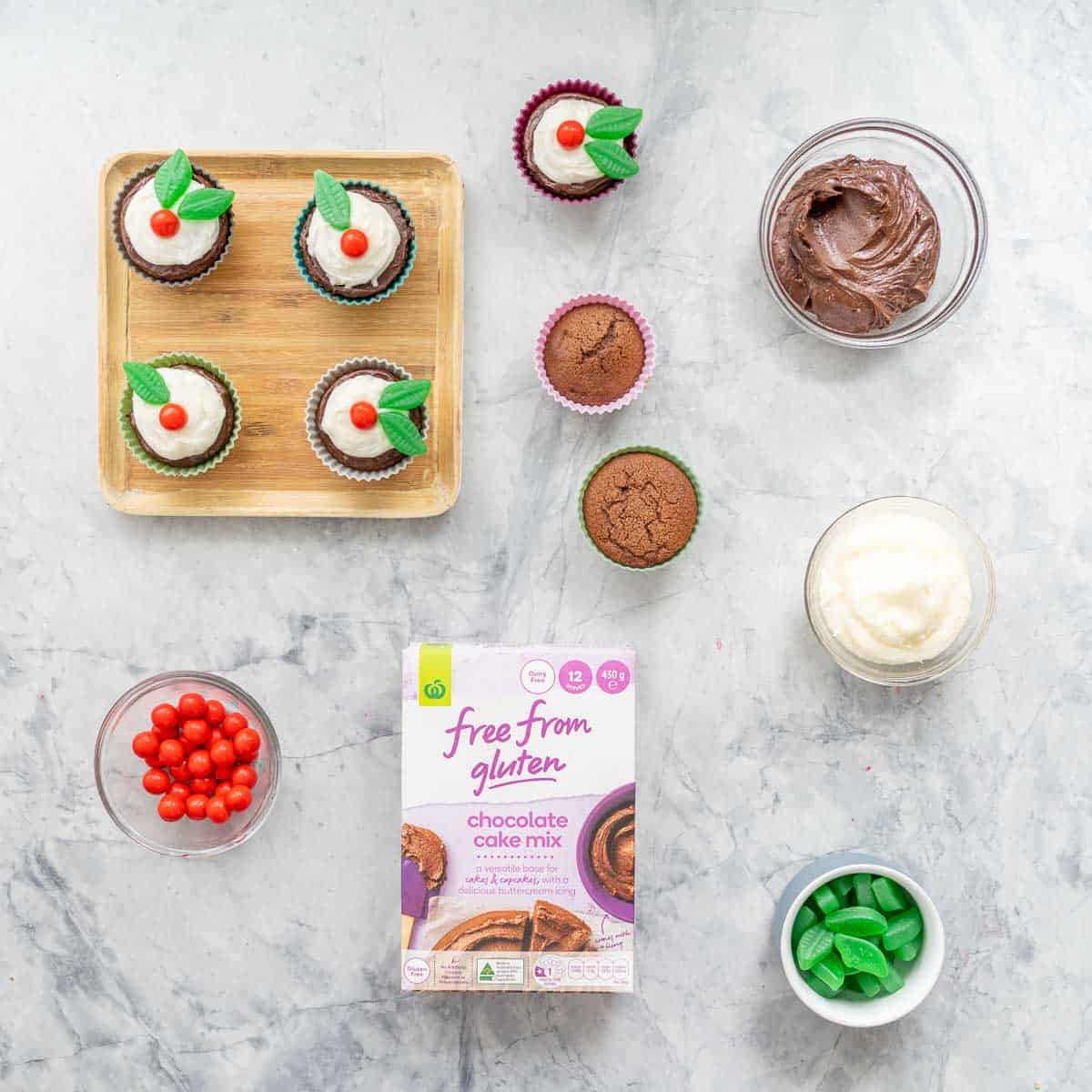 Christmas pudding decorated cupcakes, a chocolate cake packet mix, candy and bowls of frosting on a marble bench top.