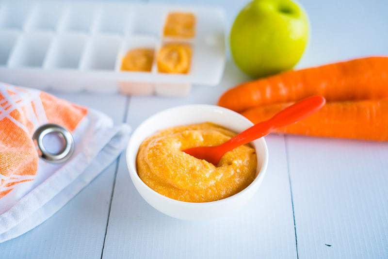 baby cereal in a a white bowl with orange spoon, in the background is the same quinoa cereal in a ice cube container for the freezer