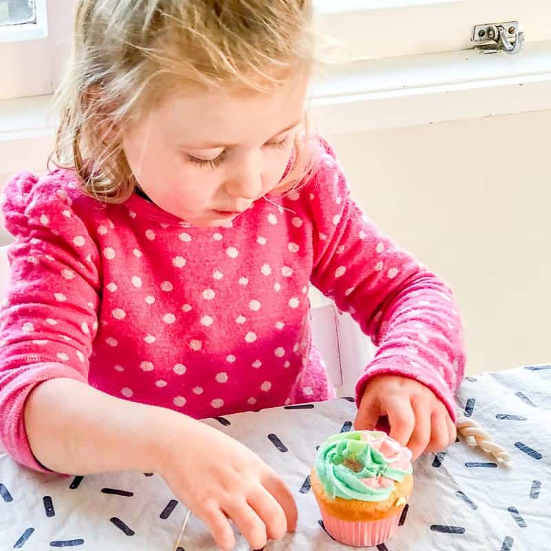 Little girl sitting at table holding unicorn cupcake.