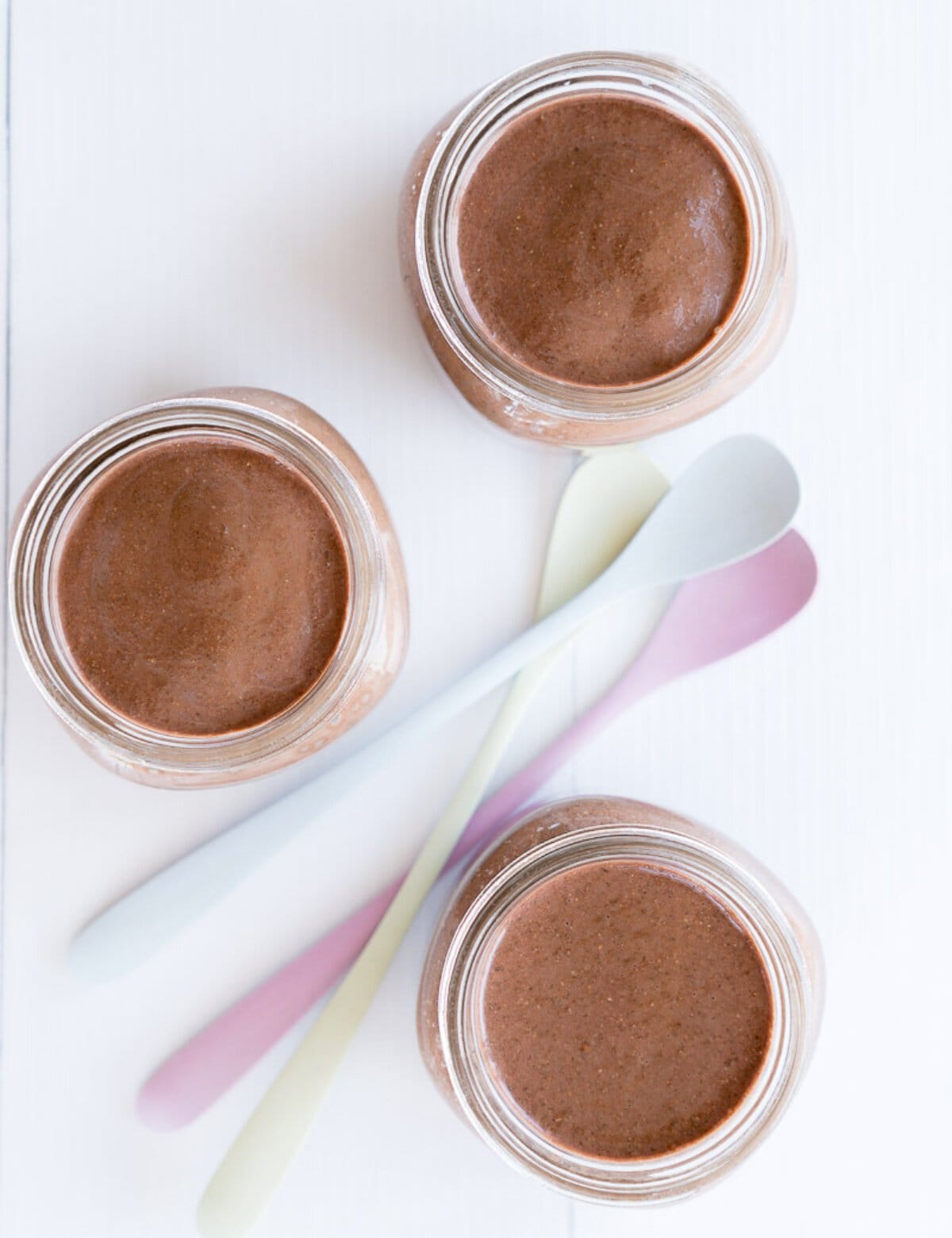 Looking down into three glass jars filled with smooth chocolate pudding next to three long handled dessert spoons.