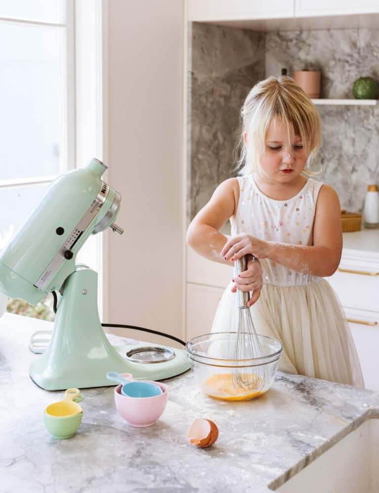A toddler in a white dress standing in a kitchen next to a floured bench top and green cake mixer.
