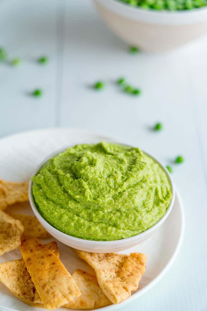 A bowl of green pea pesto next to pita crisps.
