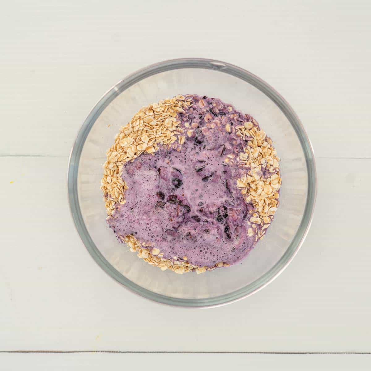 Blueberry mixture inside a large glass bowl of oats on bench top.