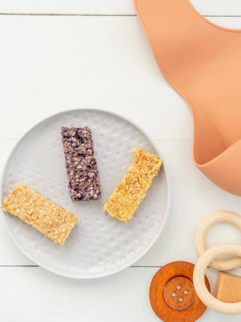 Three flavors of porridge fingers on a plate next to a chew toy and baby bib.