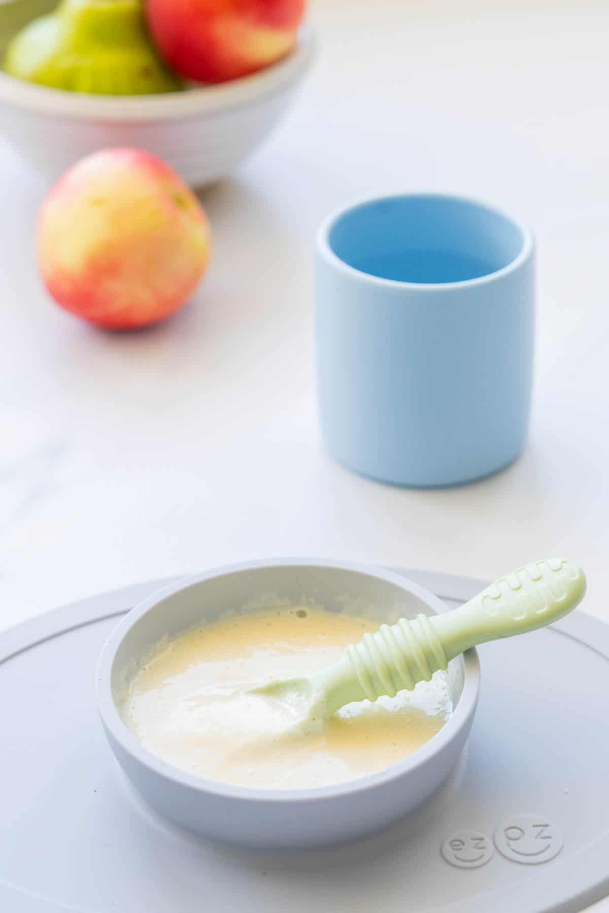 A grey silicone baby plate of baby custard with a green spoon, blue baby cup and fruit in the background.
