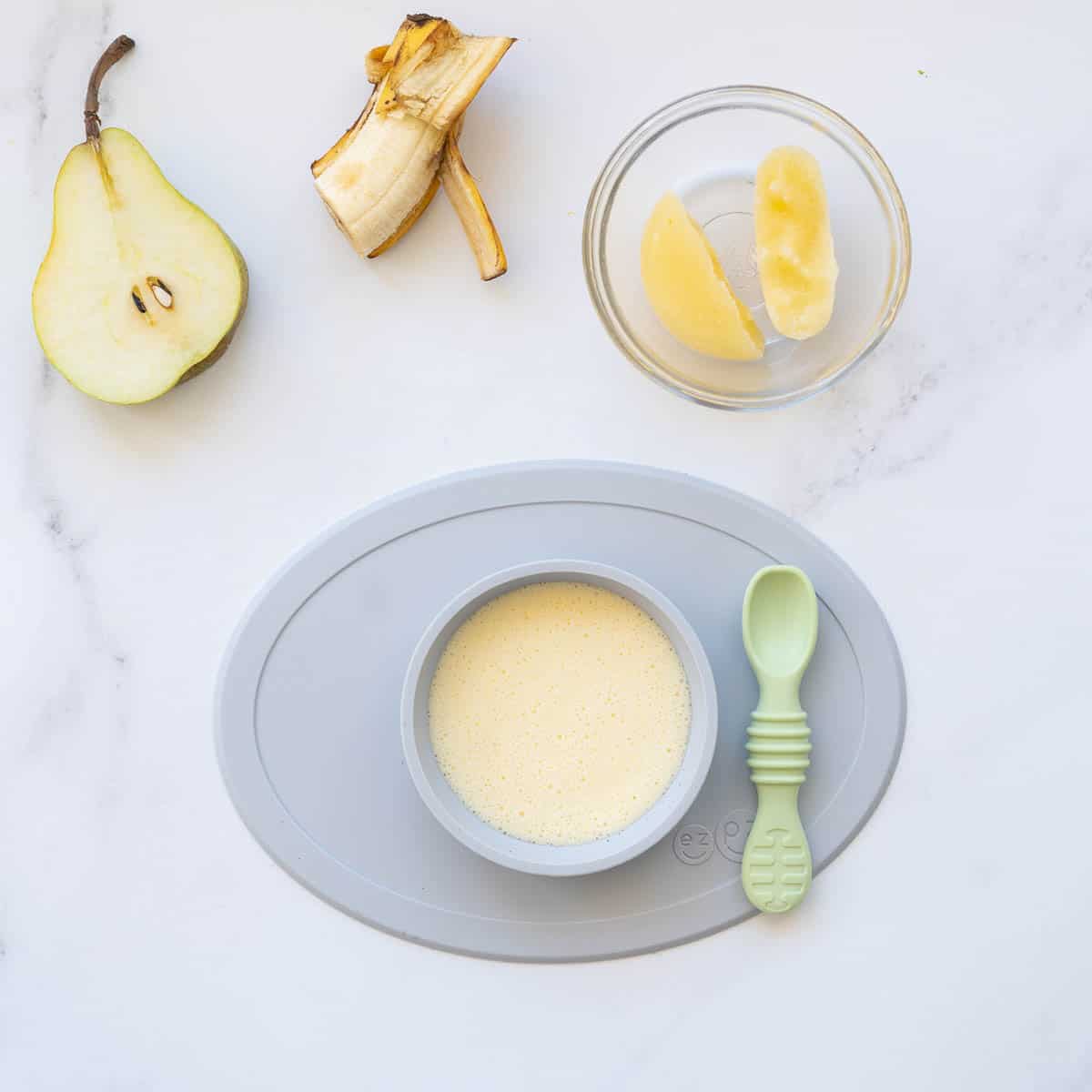 A baby plate of custard with a spoon, next to a banana, frozen fruit puree and a pear slice.