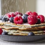 Thin pancakes stacked on a plate topped with fresh raspberries and blueberries and icing sugar being sprinkled on.