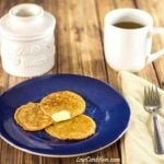 Blue plate with three small pancakes with piece of butter melting, mug of coffee, fork on table.