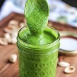 Tall glass jar on wooden board with bright green sauce inside, desert spoon dipped in and held above jar with sauce dripping from spoon.
