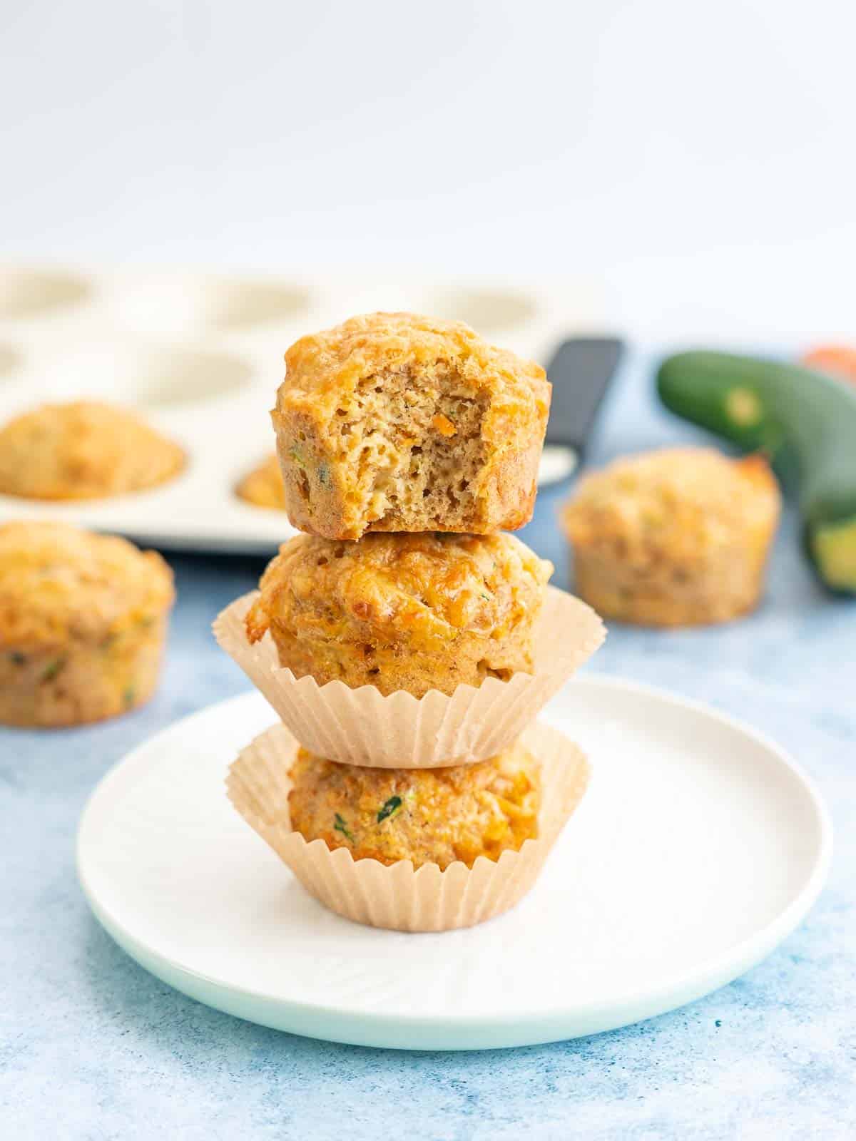 3 savoury muffins staked on top of each other, muffin tray and vegetables in the background