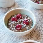 Four white bowls on wooden bench with chocolate mousse topped with coconut and raspberries.