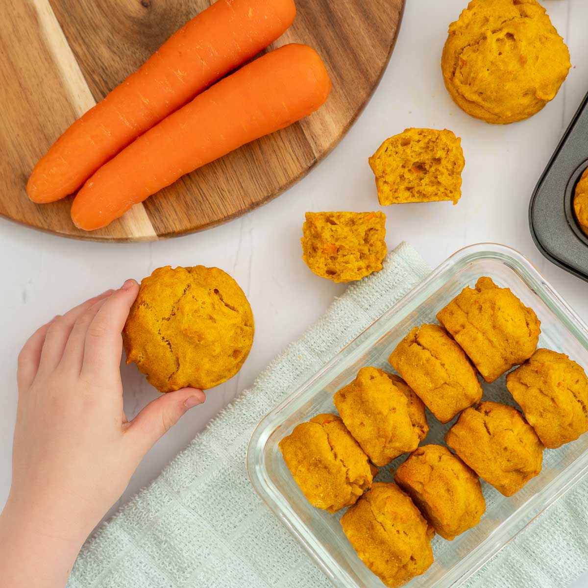 container of mini carrot muffins on a bench top with a child's hand reaching for a muffin