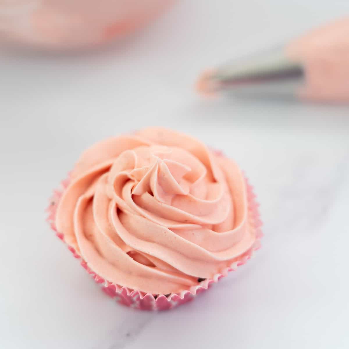 Cupcake frosted with piped pink vegan buttercream sitting on bench top.