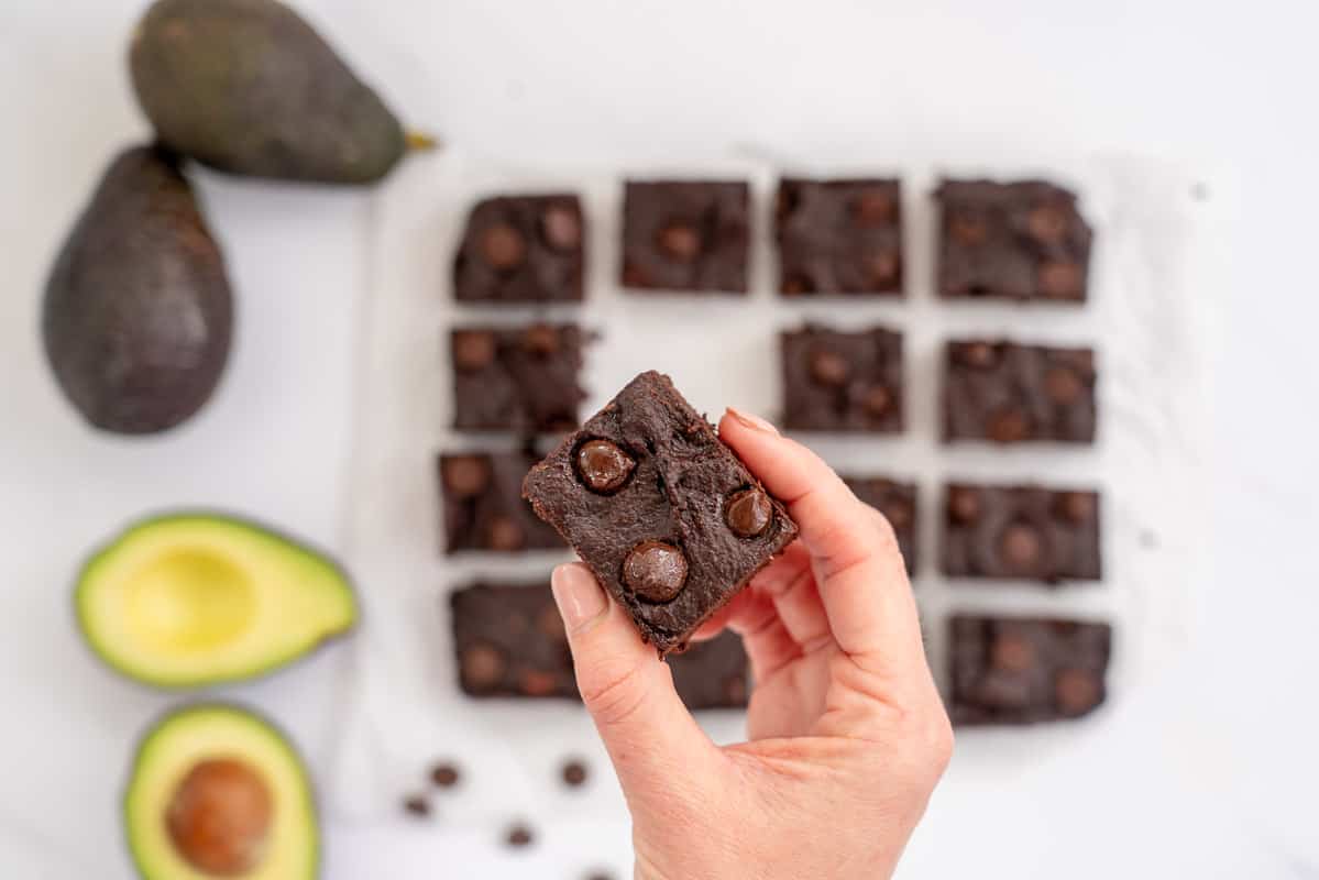 hand holding a piece of avocado brownie above a halved avocado