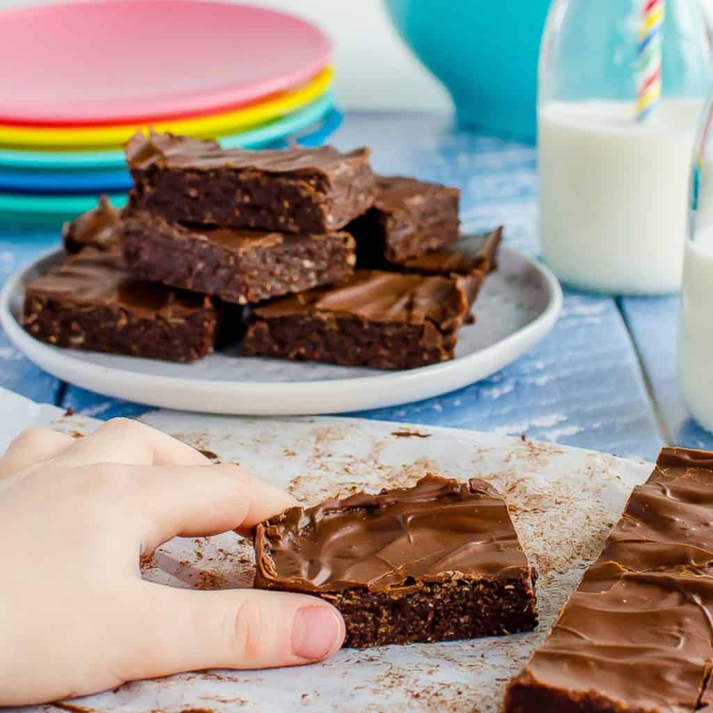 A child's hand reaching for a piece of weetbix slice sitting on a blue table top.