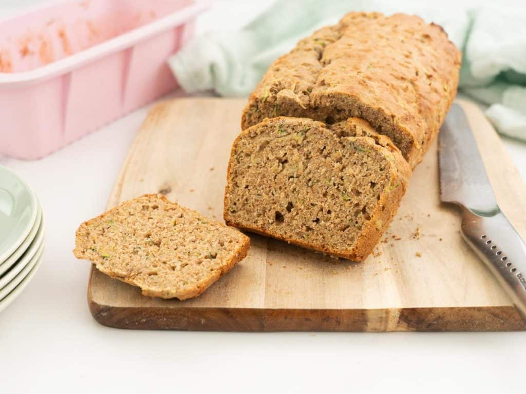 Sliced zucchini banana bread on a wooden chopping board with a pink loaf tin in the background
