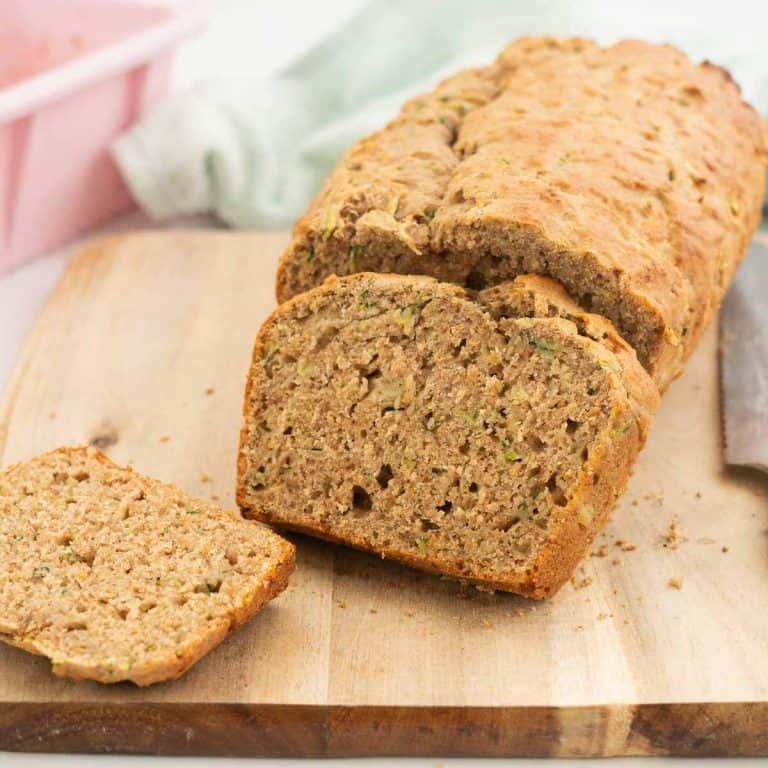 Sliced zucchini banana bread on a wooden chopping board with a pink loaf tin in the background