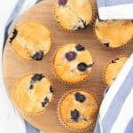 7 blueberry muffins on a round wooden chopping board, partly covered with a blue and white striped tea towel.