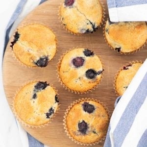 7 blueberry muffins on a round wooden chopping board, partly covered with a blue and white striped tea towel.