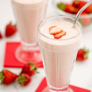 Two strawberry milkshakes in tall glasses topped with strawberry slice with red napkins.