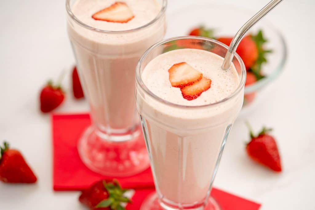 A close up of Two strawberry milkshakes in tall glasses topped with strawberry slice with red napkins.