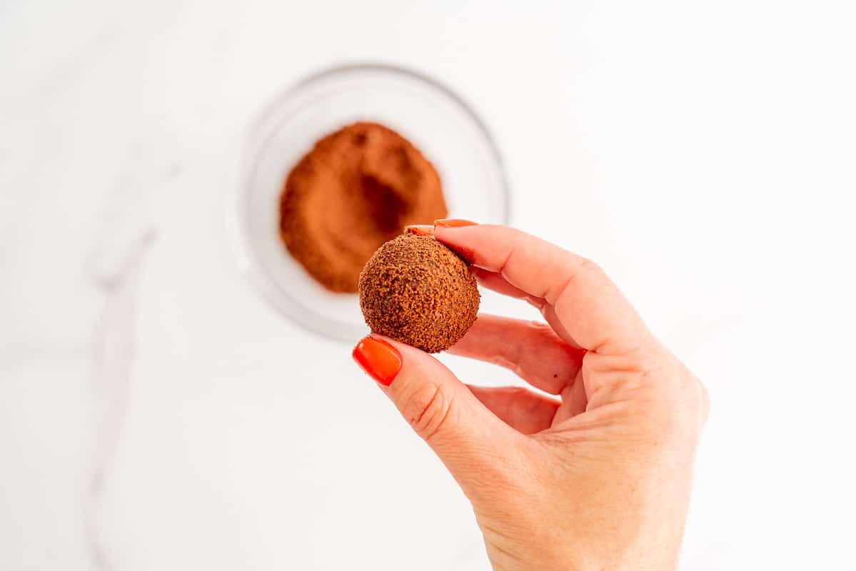 A woman's hand holding a milo coated chocolate snack ball above a small glass bowl of milo.