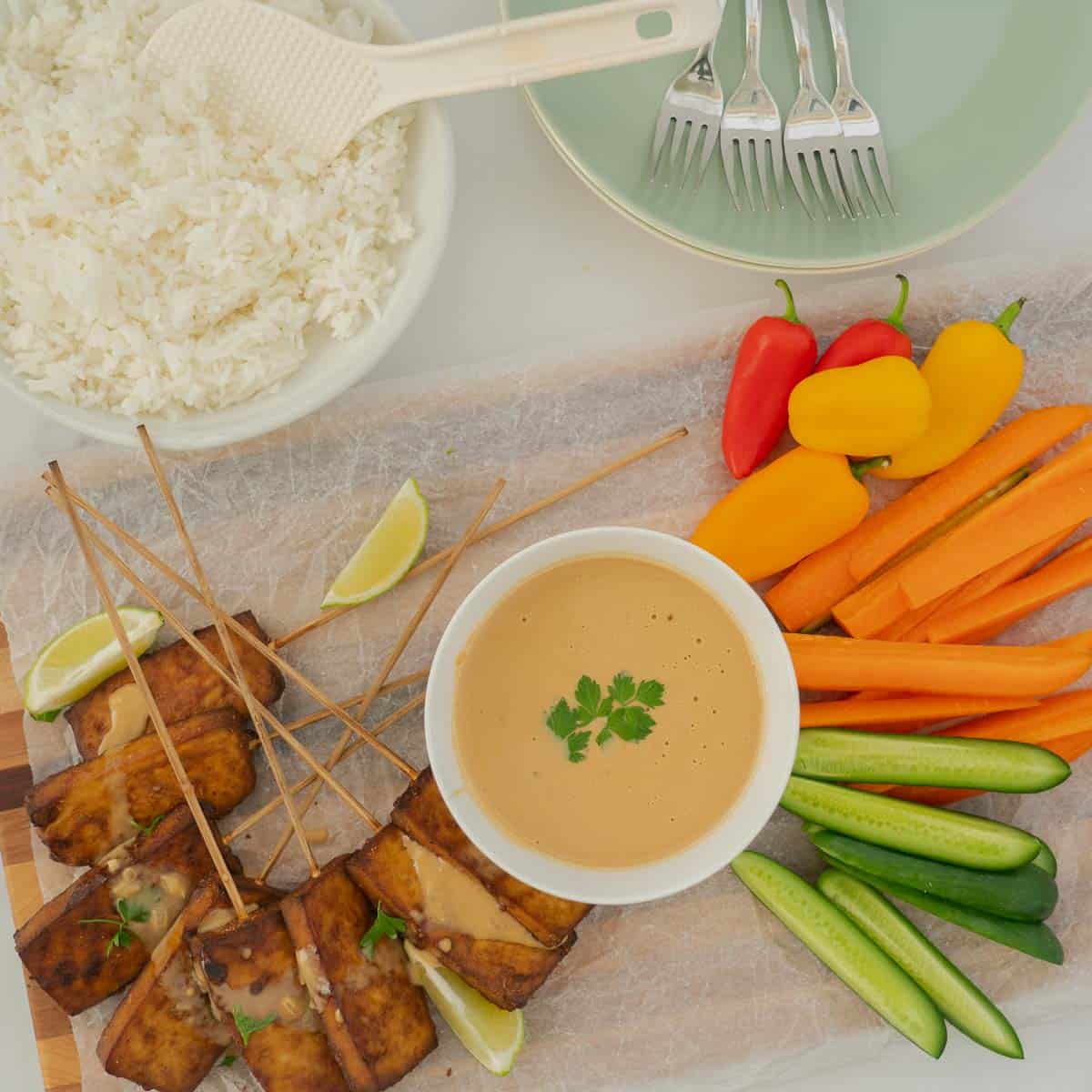 Top down photo showing baked tofu, satay sauce and vegetables laid out on a platter with a bowl of steamed rice.