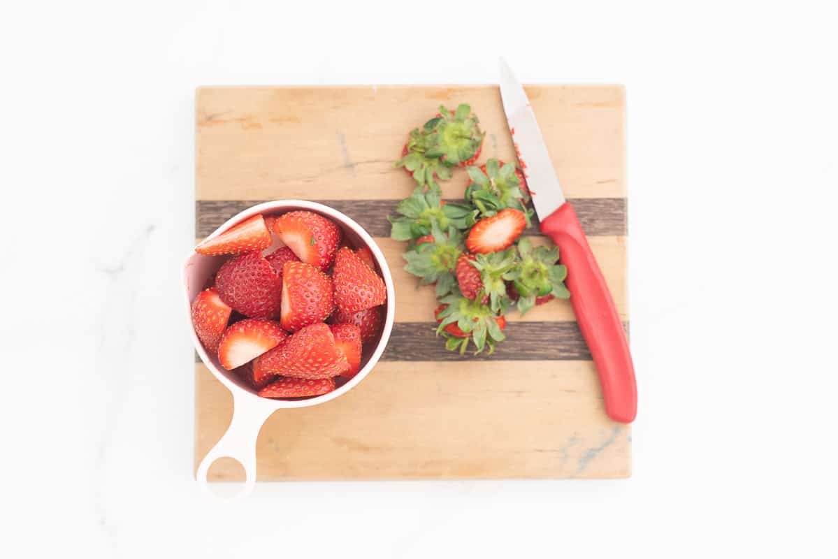 A full measuring cup of sliced strawberries nexy to a chopping board with nice and removed strawberry tops.