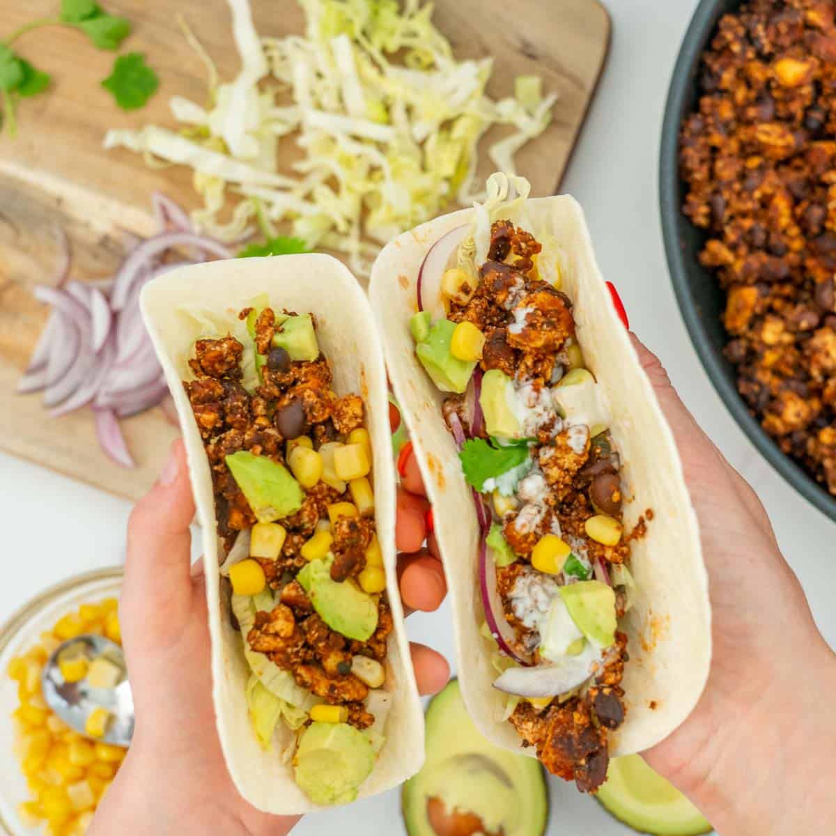 Two tacos being held up above a spread of fillings, one held by a Childs hand and one held by an adults hand.