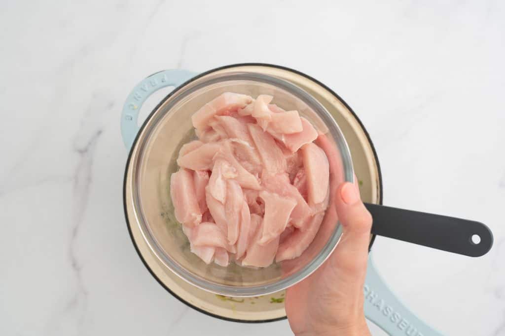 A woman's hand holding a glass bowl of sliced chicken breast above a large ceramic dish.