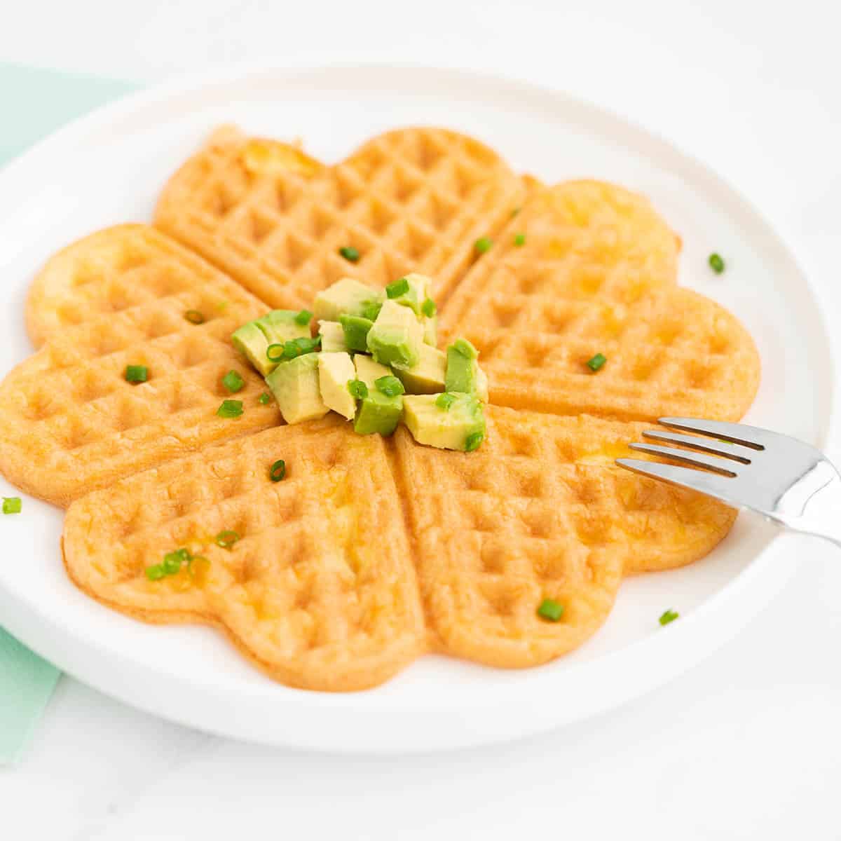 An egg waffle on a white plate garnished with small cubes of avocado and chives.