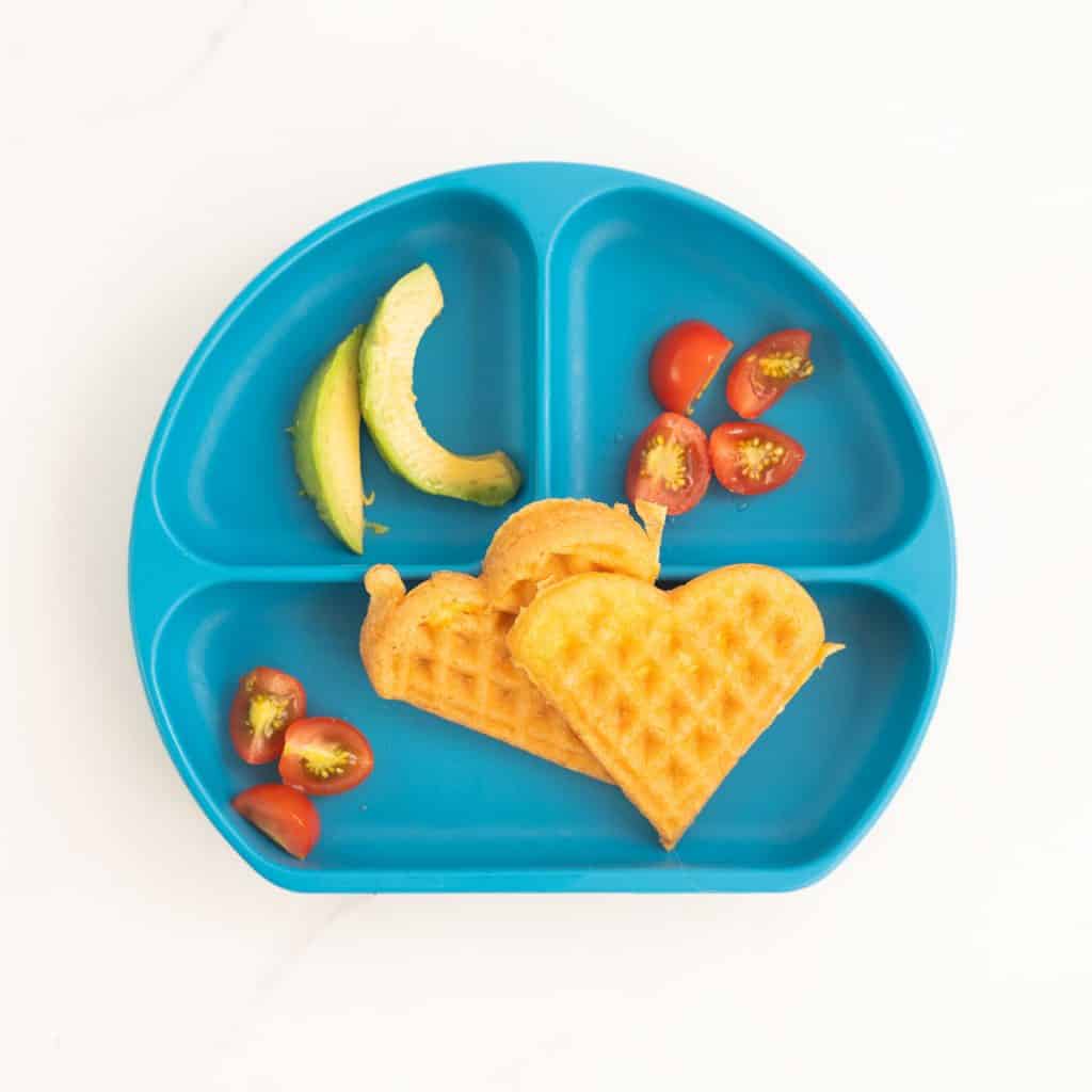a blue silicone divided plate with 2 heart shaped egg waffles, sliced tomatoes and avocado.