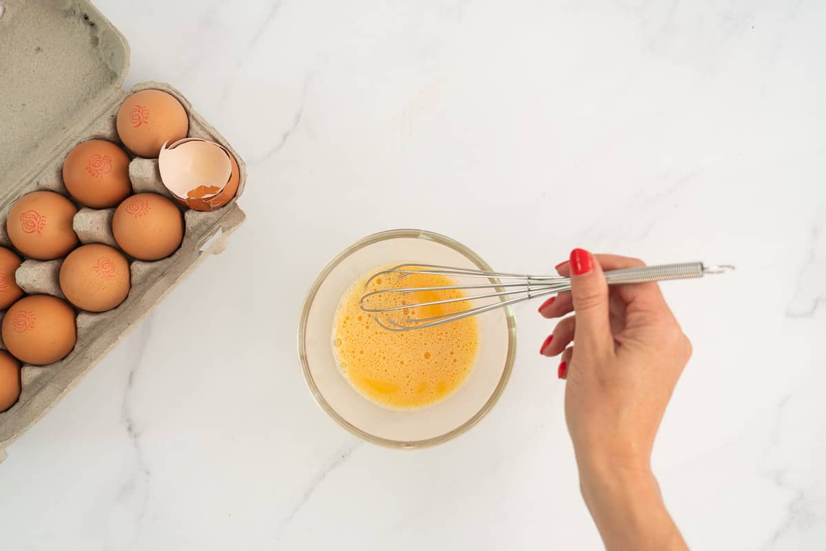 woman's hand holding a whisk above a well beaten egg, small bubbles visible on the surface of the egg.