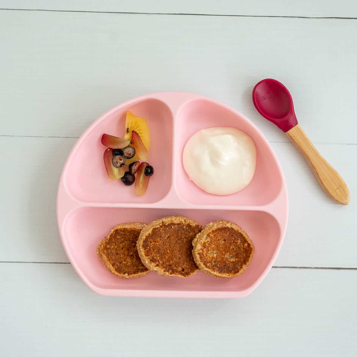 A pink divided baby plate, with 3 baby pancakes, sliced fruit and yoghurt.