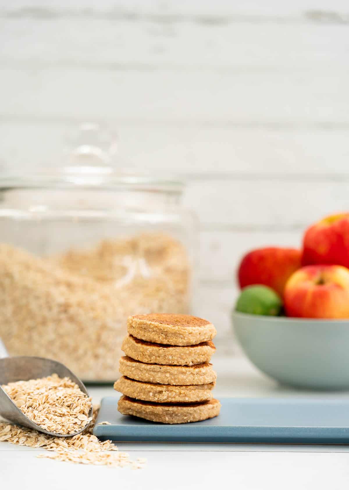 A stack of baby pancakes with a bowl of apples and jar of rolled oats in the background