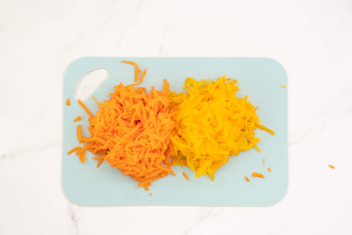 Piles of grated carrot and grated pumpkin on a blue chopping board.