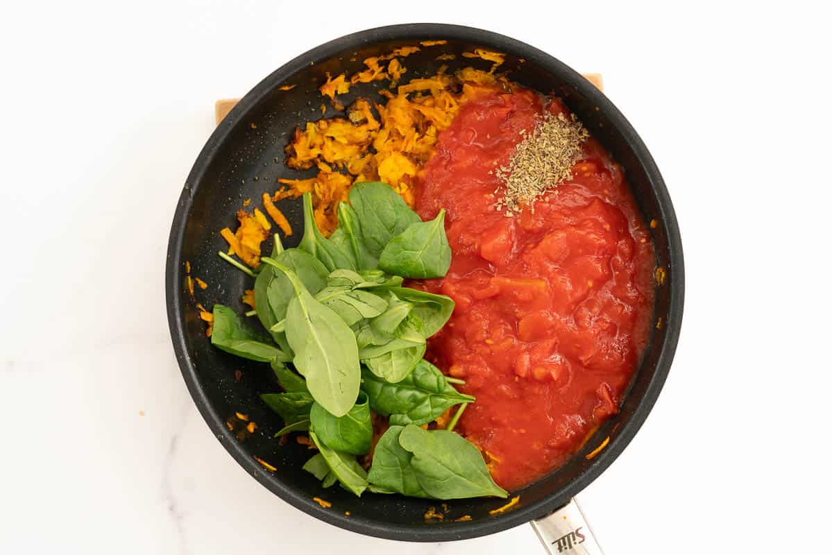 Cooked grated carrot and pumpkin, with tinned tomatoes, dried herbs and spinach leaves in a fry pan.